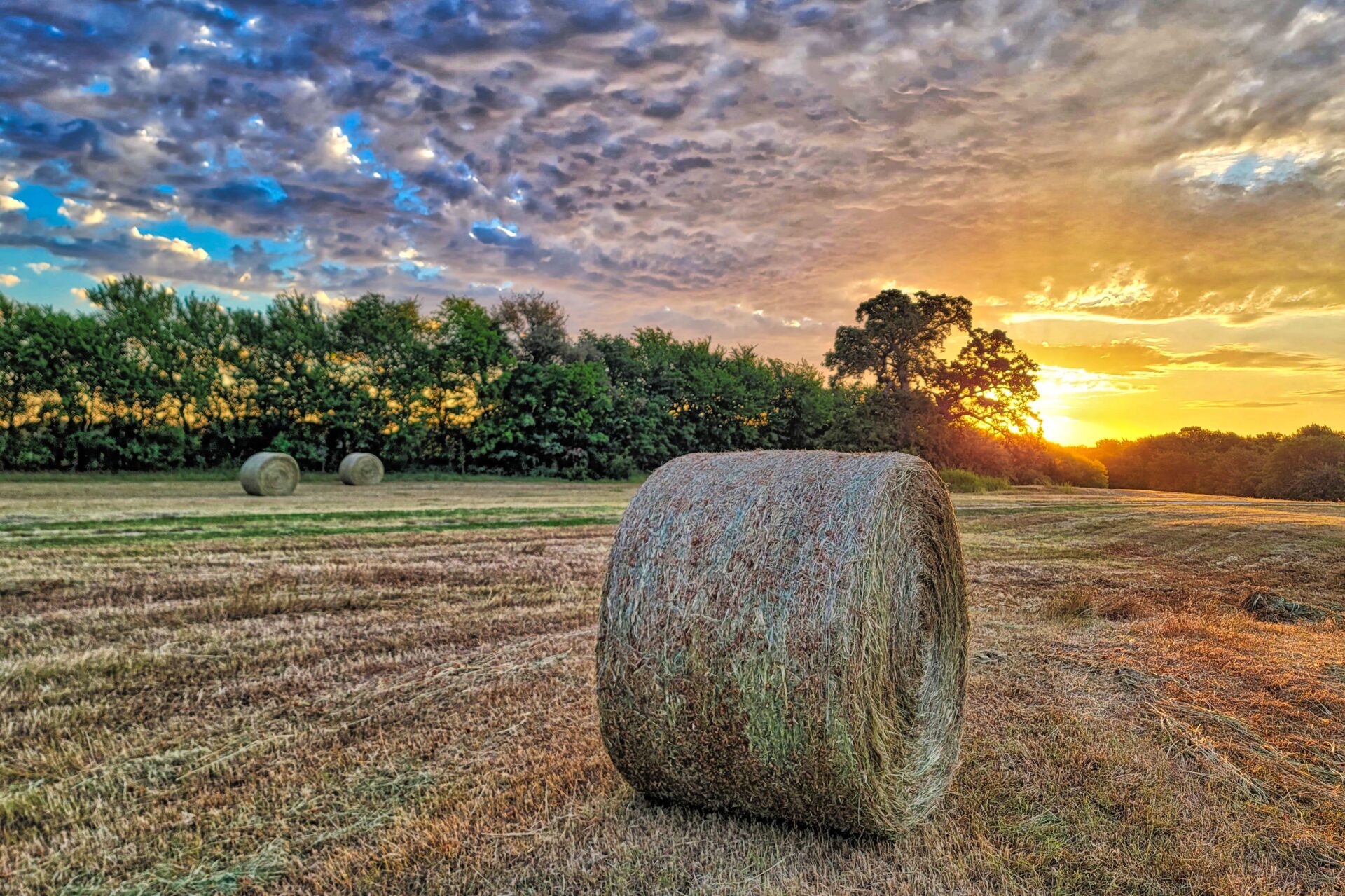 Round hay bale in a field with a bright sunset peaking through clouds.