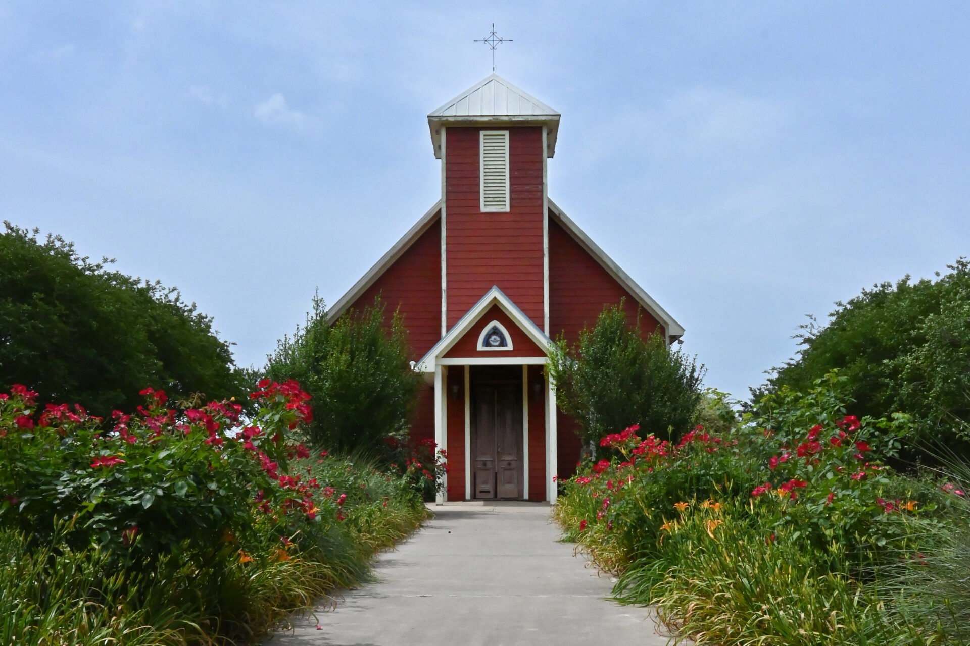 Red rural church surrounded by bright green plants, blooming flowers, and a blue sky.