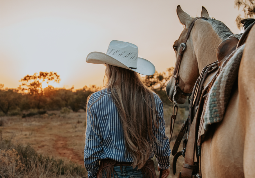 Blonde woman in a cowboy hat facing away with a horse saddled.
